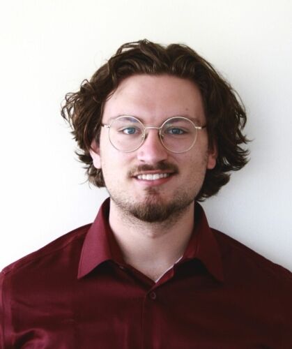 Young man with curly brown hair, glasses, and a friendly smile, wearing a maroon shirt.