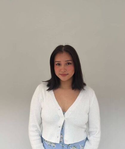 Young woman with dark hair wearing a white cardigan and floral dress, standing against a plain light grey background, representing young Canadians advocating for resource development and sustainable growth.