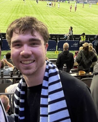 Young man at a soccer match smiling, wearing a striped scarf, with the stadium and fans in the background.