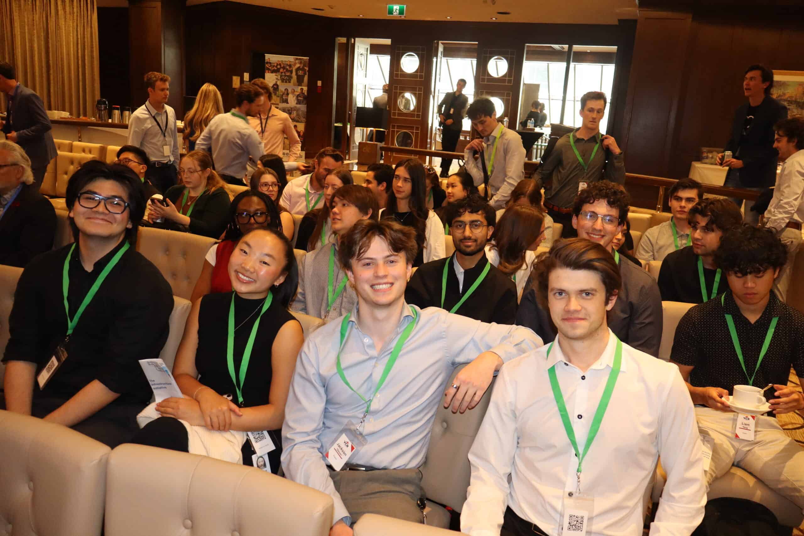 AI-generated group photo of young professionals attending a conference, featuring diverse individuals wearing ID badges in a warm, indoor setting filled with people, indicating a formal networking or seminar event.