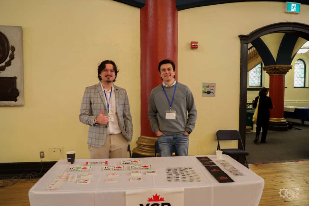 Two young professionals at a chemical engineering career fair, standing behind a table with informational materials, in a historic building with arched windows.