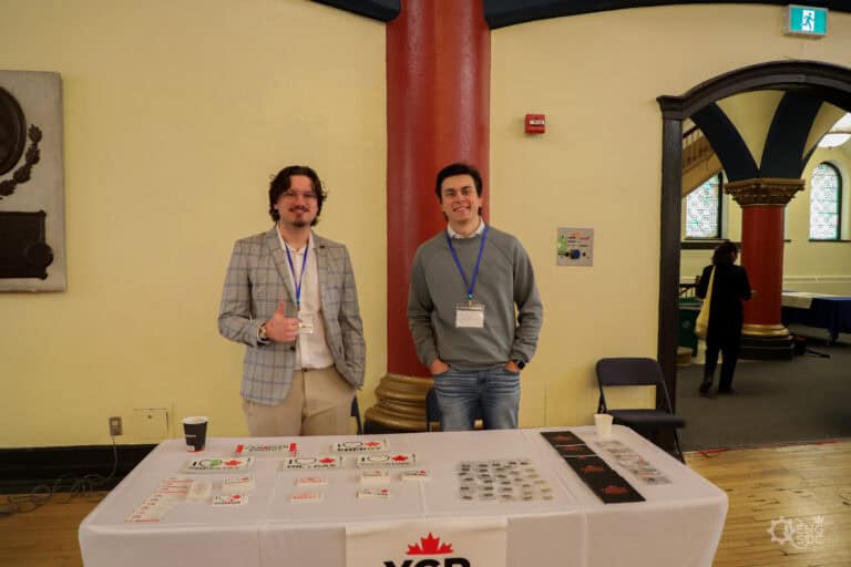 Two young professionals at a chemical engineering career fair, standing behind a table with informational materials, in a historic building with arched windows.