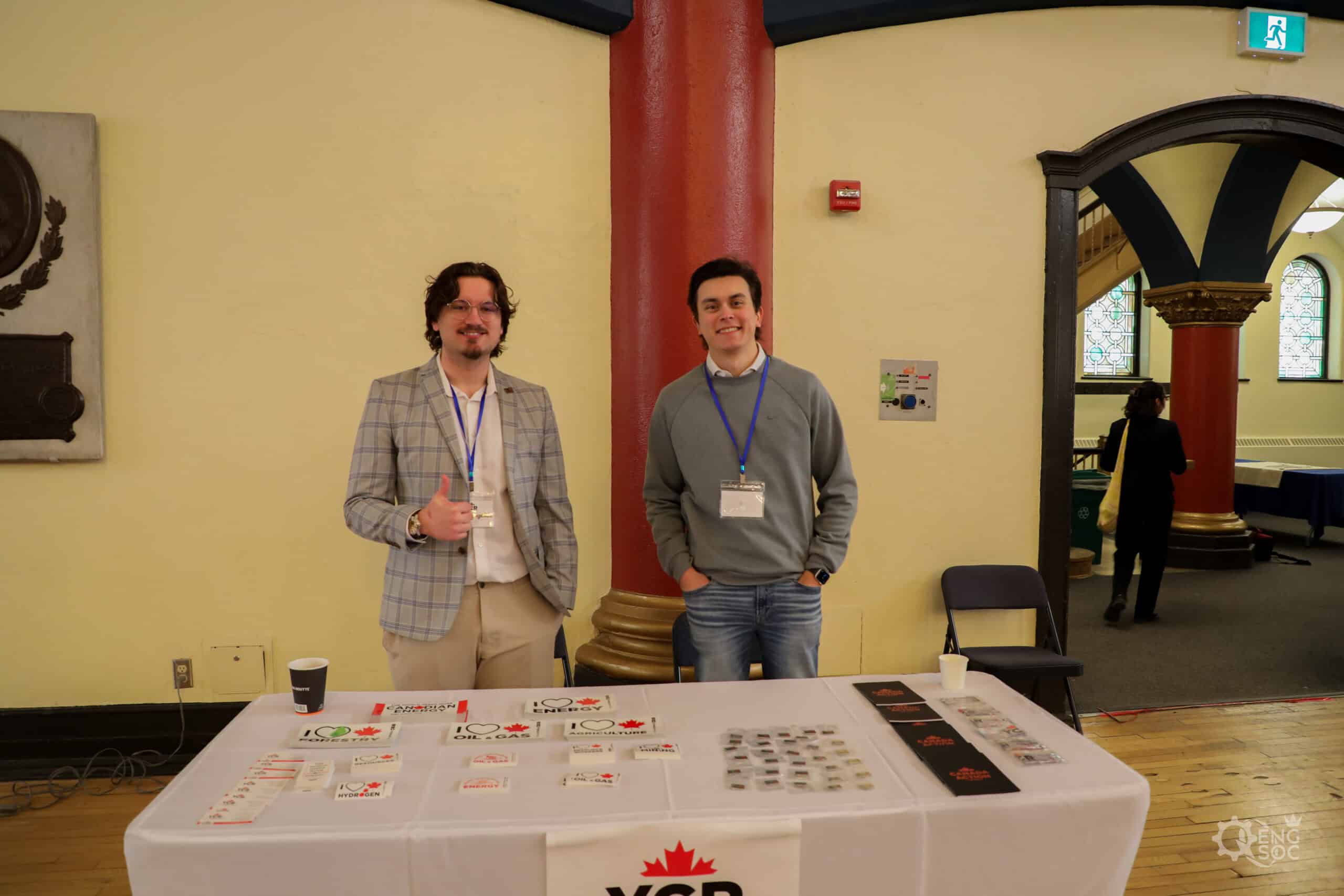 Two young professionals at a chemical engineering career fair, standing behind a table with informational materials, in a historic building with arched windows.