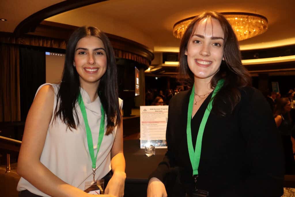 A woman with long dark hair and a white sleeveless top, and another with shoulder-length brown hair and a black blazer, both wearing green lanyards, smiling at a professional conference.
