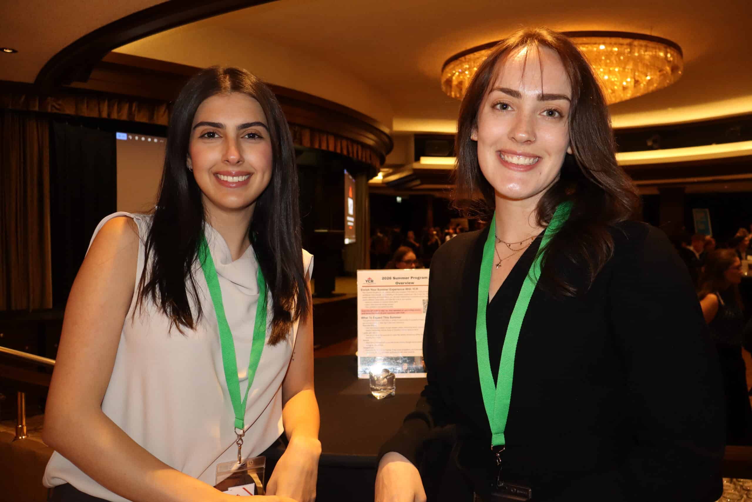 A woman with long dark hair and a white sleeveless top, and another with shoulder-length brown hair and a black blazer, both wearing green lanyards, smiling at a professional conference.