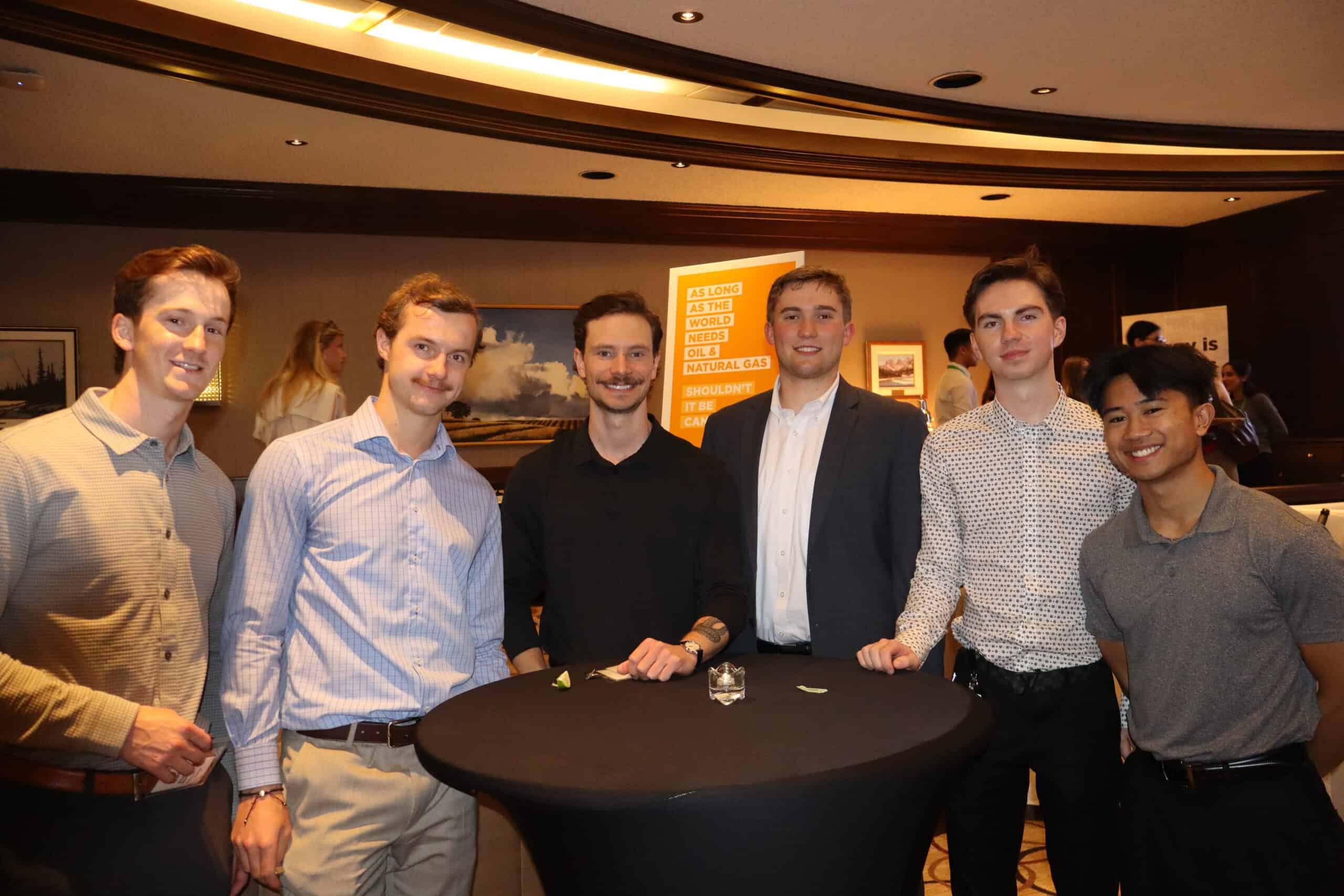Group of six diverse young men smiling and standing around a high-top table at a professional networking event.