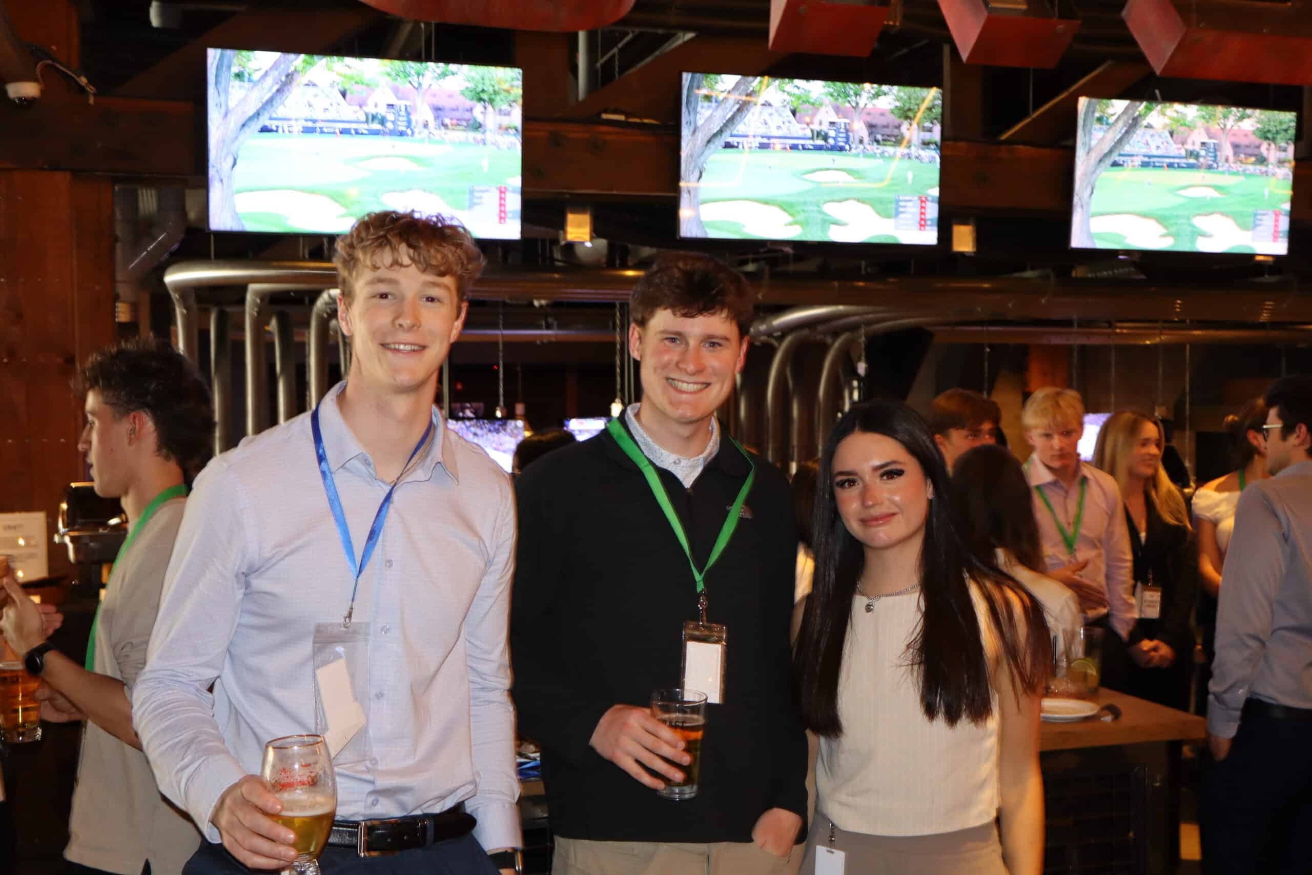 A group of three young adults, two men and one woman, smiling and holding drinks at a social gathering in a bar with multiple screens showing sports events.