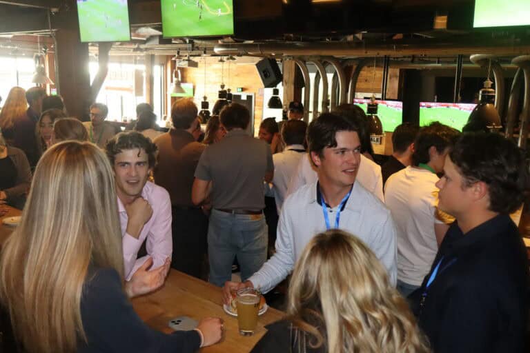 Group of young adults enjoying drinks and watching sports on TV screens in a lively bar setting.